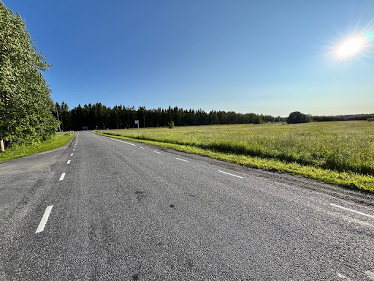Asfaltová silnice vinoucí se krajinou kolem zelených polí a lesa, slunečný letní den s modrou oblohou.