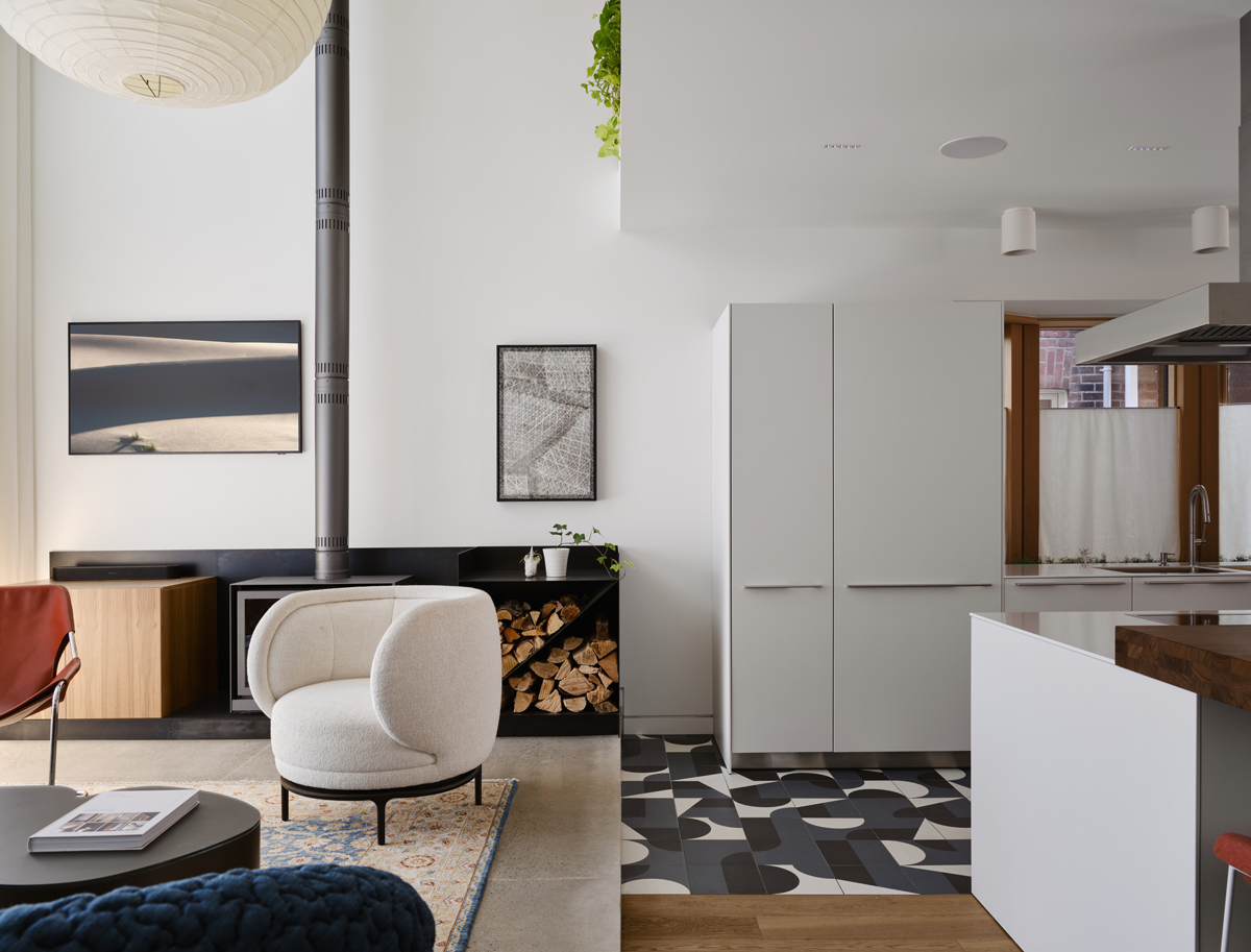 Světlý obývací pokoj s bílým designovým křeslem, krbovými kamny a kuchyní s originální dlažbou.
