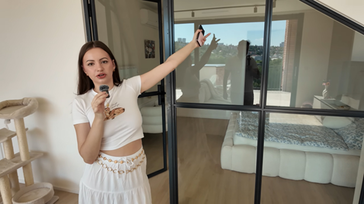 Mladá žena v bílé sukni ukazuje na prosklenou stěnu s černými rámy v moderním bytě, za ní je vidět ložnice s nízkou postelí a výhledem na terasu.