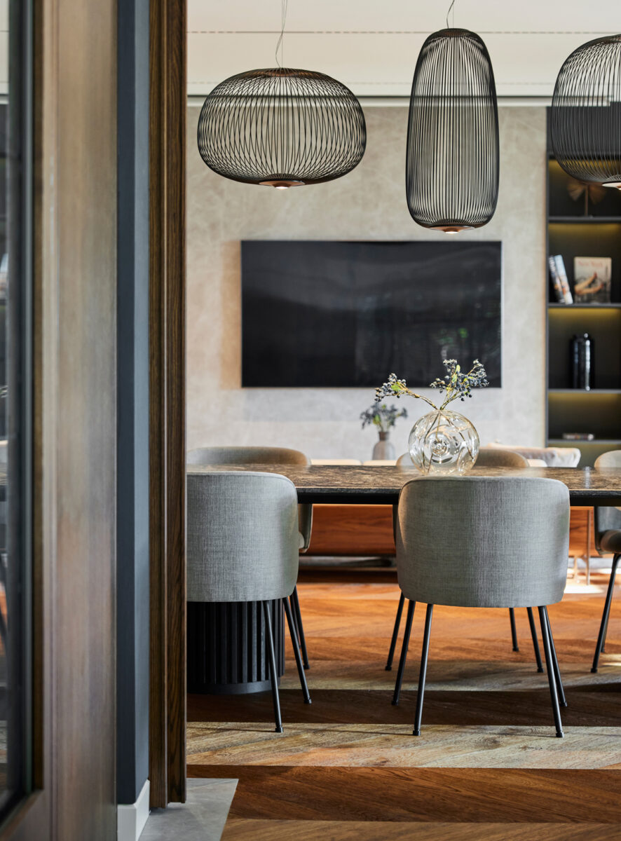 Detailní pohled na elegantní čalouněné židle v jídelně, doplněné originálními designovými svítidly z černého kovu, které tvoří výrazný kontrast k jemné kresbě dřevěné podlahy.