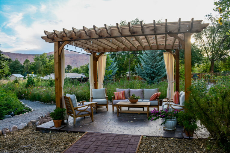 Dřevěná pergola s pohodlným venkovním posezením, polštáři a závěsy uprostřed rozkvetlé zahrady.