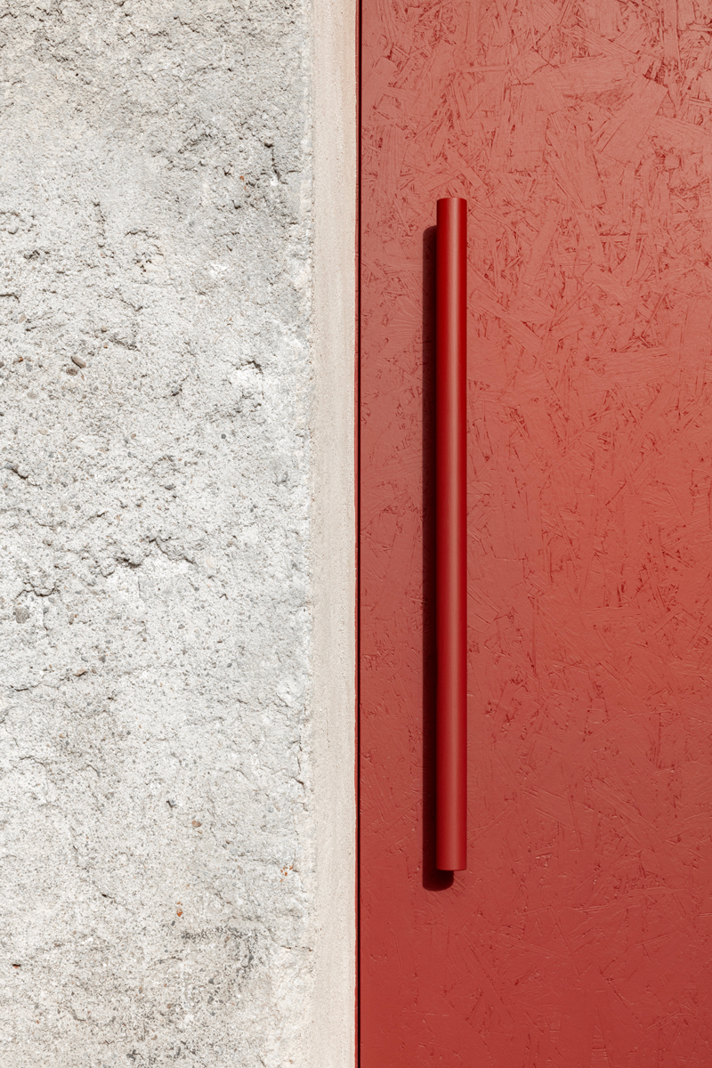 Minimalistický detail matně červených dveří s výrazným madlem, které láká k otevření a vstupu do klidného prostoru uvnitř.