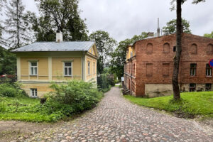 Úzká dlážděná ulička mezi žlutým dřevěným domem a cihlovou budovou, lemovaná stromy a keři v malé estonské vesnici.