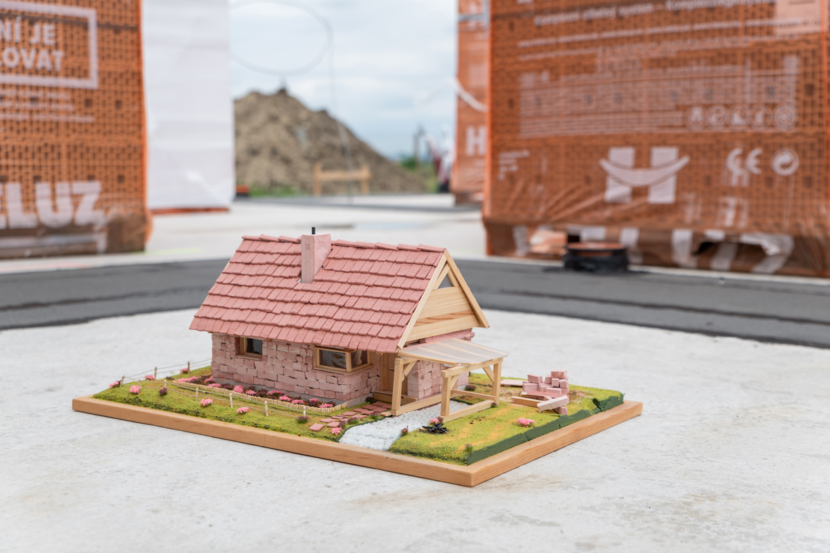 Detailní záběr na model rodinného domu postaveného z miniatur cihel, umístěný na betonové desce stavby, v pozadí balíky cihel HELUZ.