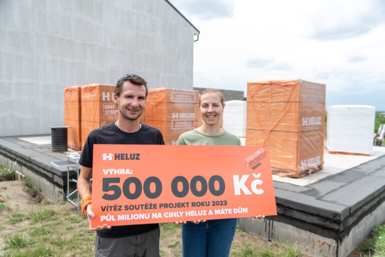 Dvojice držící velký oranžový šek s výhrou 500 000 Kč v soutěži HELUZ Projekt roku 2023, na stavbě s balíky cihel v pozadí.