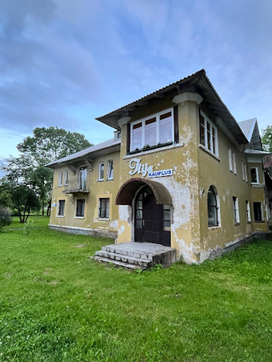 Starý žlutý dům se zašlou omítkou a nápisem „Kauplus“ u vchodu, v letním zeleném parku pod modrou oblohou.