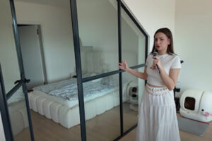 Moderní ložnice oddělená černou skleněnou příčkou, žena ukazuje do prostoru.