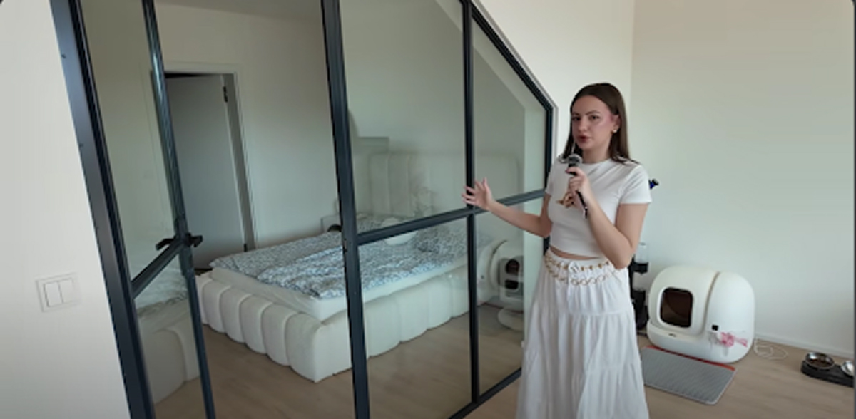Moderní ložnice oddělená černou skleněnou příčkou, žena ukazuje do prostoru.