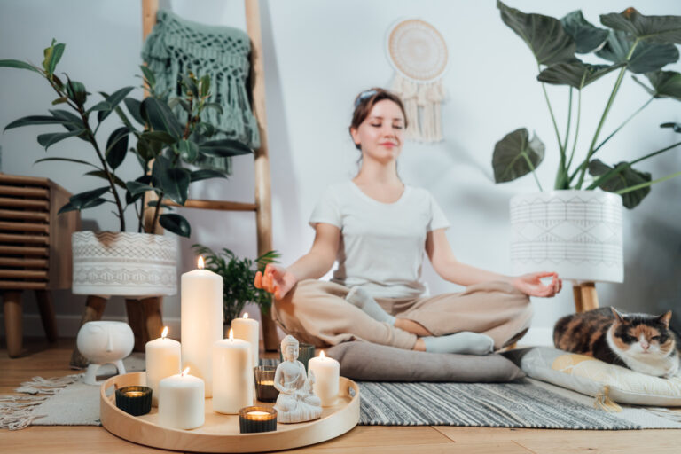 Žena sedí v meditační pozici u zapálených svíček, vedle ní leží spokojená kočka v rostlinně dekorovaném interiéru.