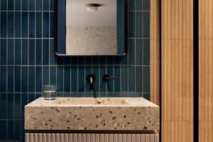 Elegantní koupelna s tmavě modrými obklady, terrazzo umyvadlem a dřevěným obložením, kombinace luxusu a přírodních materiálů.