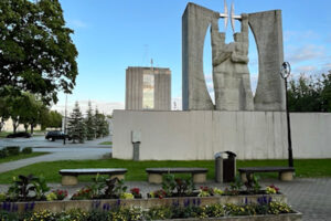 Monumentální betonový památník se sochou a květinovým záhonem na náměstí, v pozadí administrativní budova a ranní nebe.