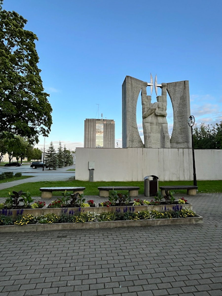 Monumentální betonový památník se sochou a květinovým záhonem na náměstí, v pozadí administrativní budova a ranní nebe.