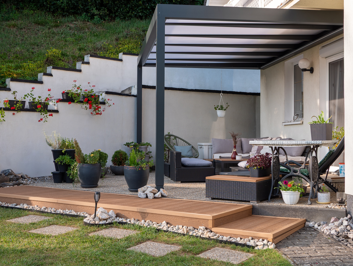 Moderní hliníková pergola s pohodlným zahradním posezením u domu, doplněná květinami v květináčích a kamennými schody.