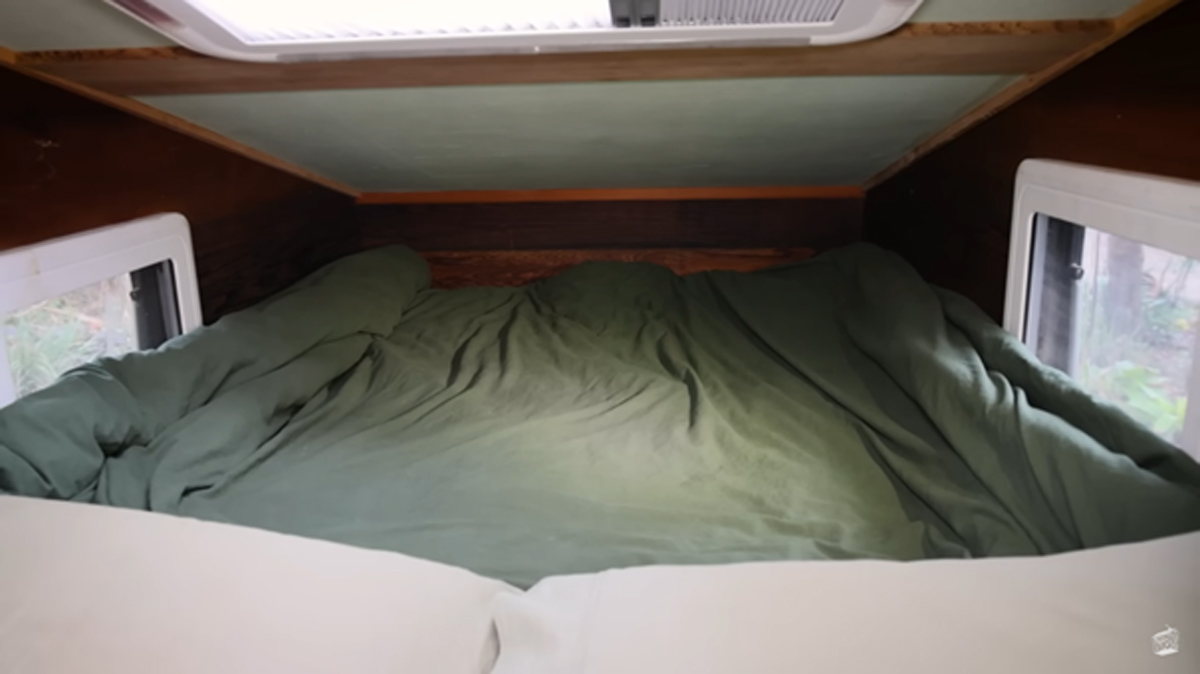 Pohled na postel s olivově zeleným povlečením pod šikmým stropem a střešním oknem v dřevěném domku. Minimalistické a útulné místo ke spaní, které evokuje klid a soukromí mezi stromy.