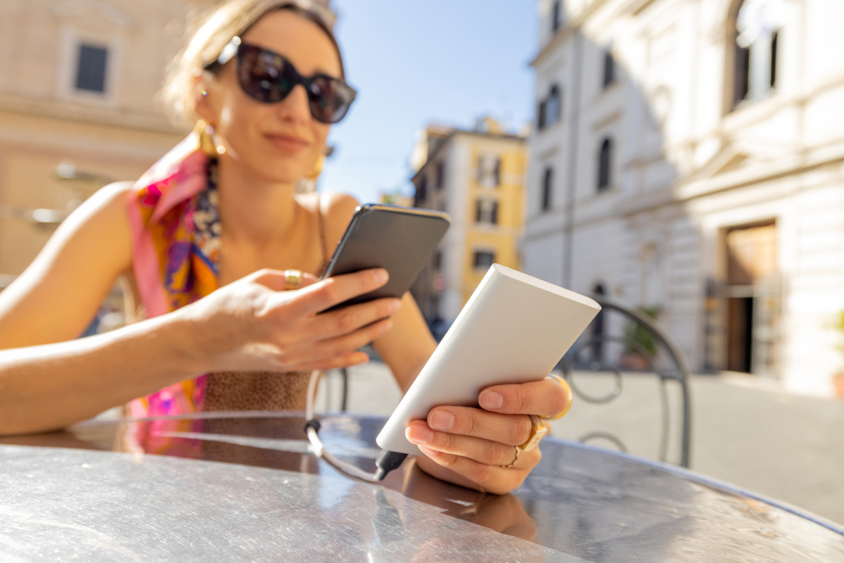 Žena v letním oblečení sedí u stolku venku a nabíjí svůj mobilní telefon z powerbanky.