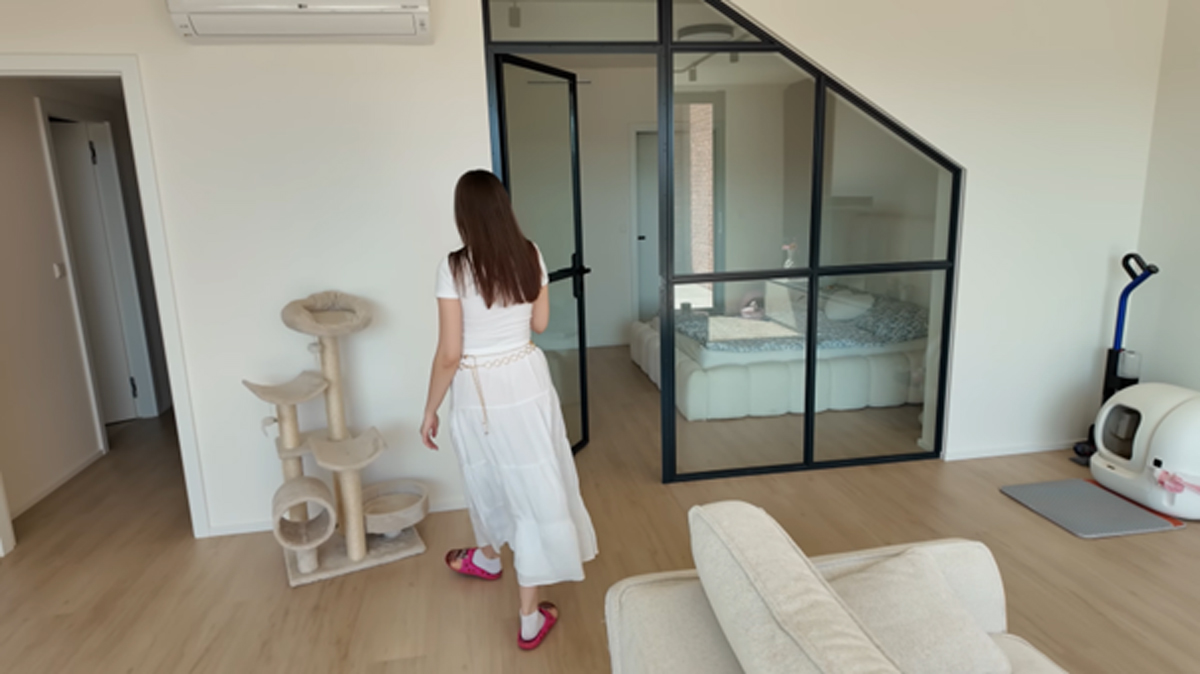 Pohled na ženu odcházející směrem k ložnici s velkou prosklenou stěnou, v popředí krémová sedačka, vlevo škrabadlo pro kočky a vpravo designový kočičí záchod. Titulek: