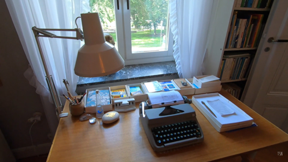 Psací stůl s retro psacím strojem, lampou a pořadači na papíry u okna s bílými záclonami, výhled do zeleně.