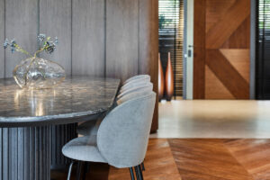 Detail oválného jídelního stolu s žilkami na desce a designových čalouněných židlí na černých nohách, v interiéru s dřevěnou podlahou.