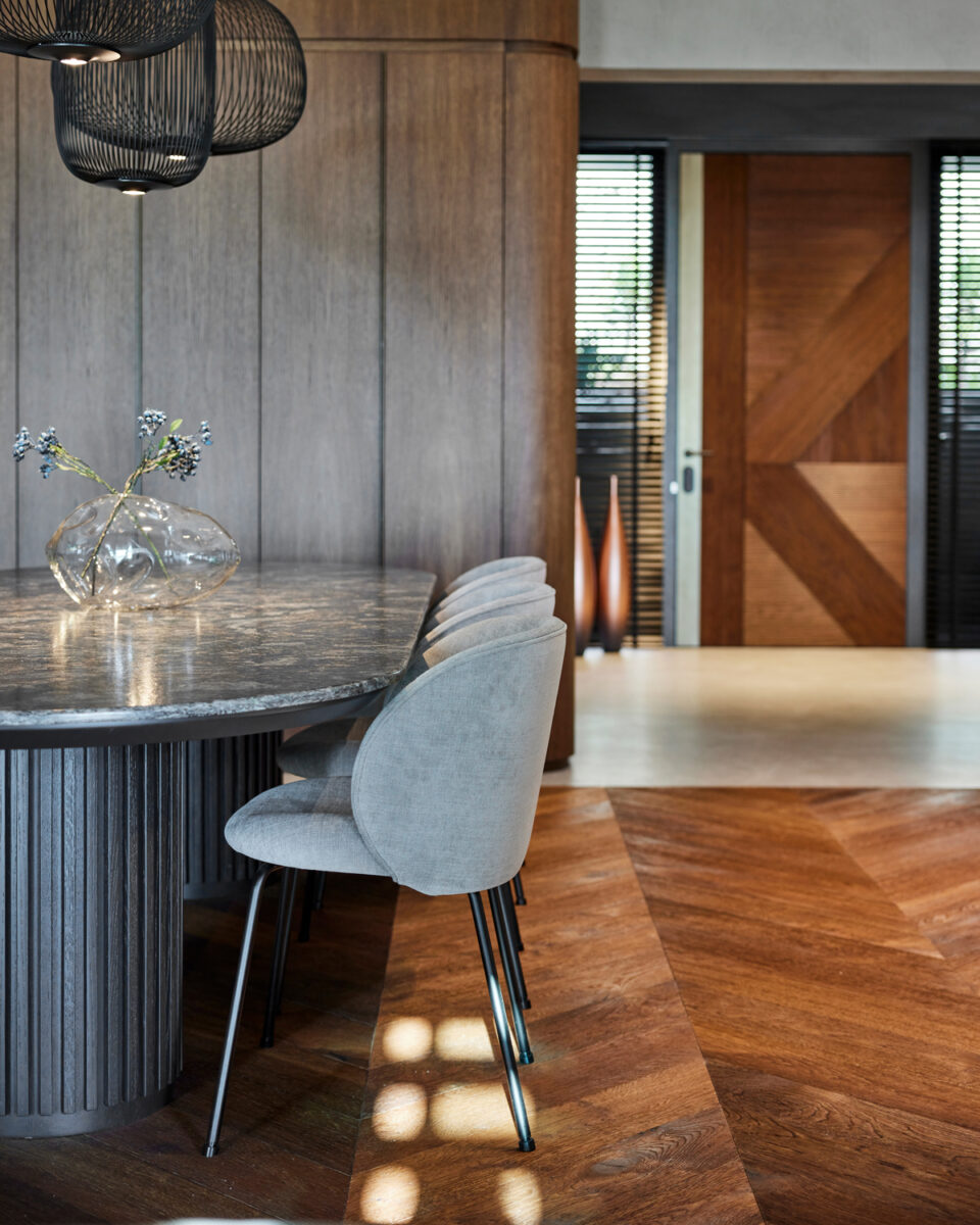 Detail oválného jídelního stolu s žilkami na desce a designových čalouněných židlí na černých nohách, v interiéru s dřevěnou podlahou.