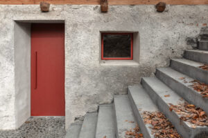 Detail vstupu s červenými dveřmi a betonovým schodištěm, kde se hrubá textura setkává s precizní linií.