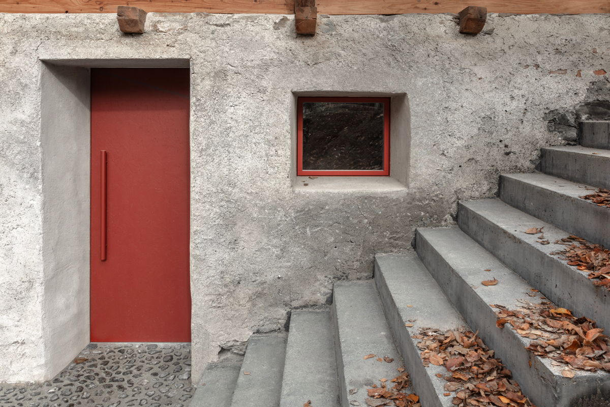 Detail vstupu s červenými dveřmi a betonovým schodištěm, kde se hrubá textura setkává s precizní linií.