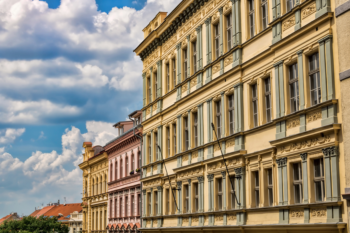  Fasády starých činžovních domů s bohatou architekturou a vysokými okny, modrá obloha s mraky v pozadí.