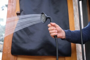 Detailní záběr na ruční sprchovou hlavici v exteriéru, z níž proudí voda. Praktický detail jednoduchého a funkčního řešení hygieny v přírodě, ideální pro život na cestách.