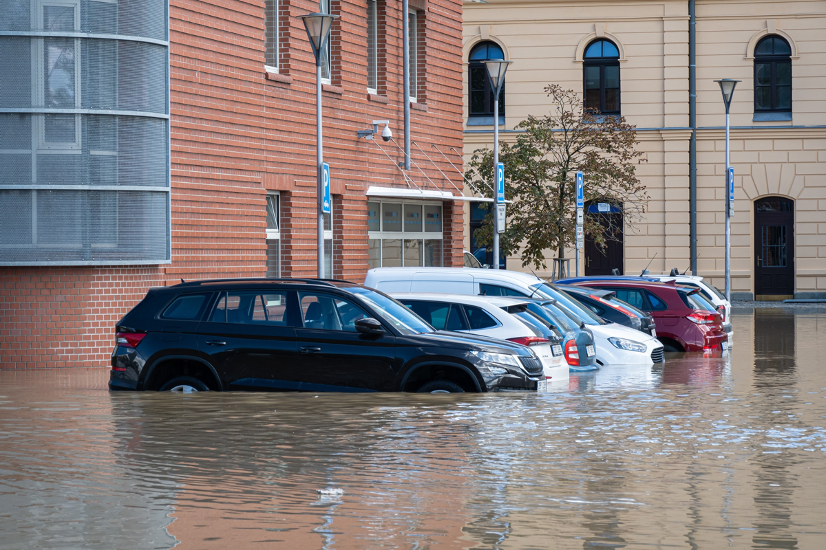 Zaparkovaná auta částečně ponořená ve vodě během povodně ve městě, v pozadí jsou budovy.