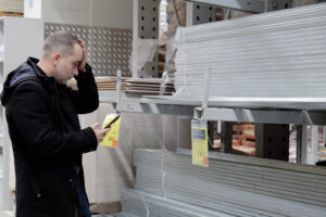 Muž v hobbymarketu sleduje telefon a nevěřícně si prohlíží rostoucí ceny materiálu.