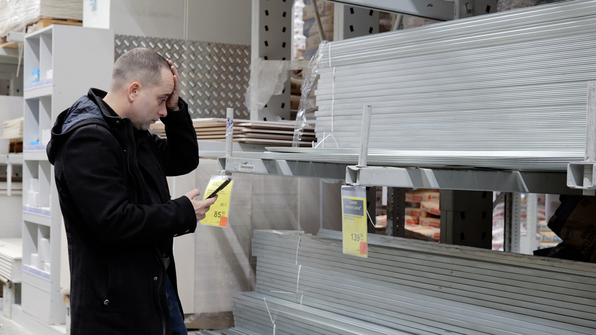 Muž v hobbymarketu sleduje telefon a nevěřícně si prohlíží rostoucí ceny materiálu.