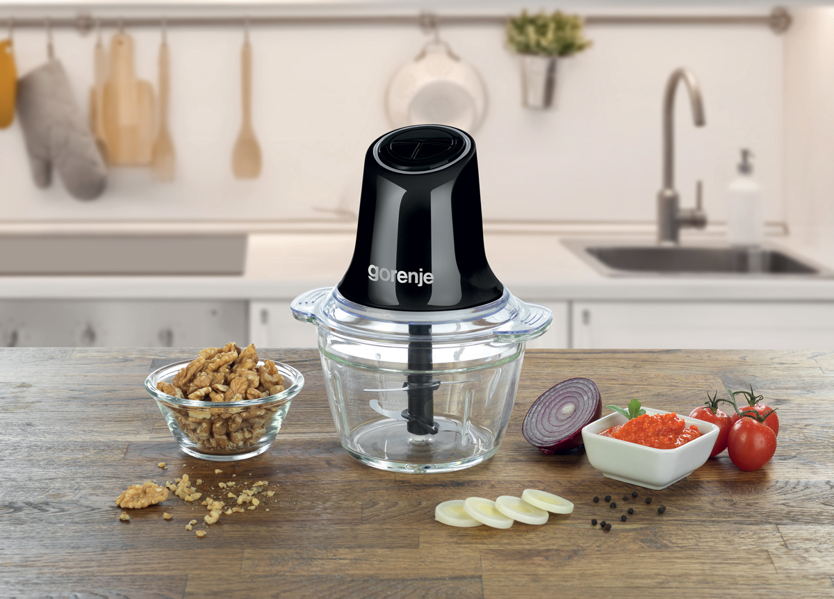 Stolní sekáček Gorenje se skleněnou mísou na dřevěné desce, kolem vlašské ořechy, plátky česneku, půlka červené cibule, rajčata a miska rajčatové salsy; v pozadí rozostřená kuchyň se dřezem.