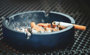 Popelník plný nedopalků s hořící cigaretou položenou na okraji stolku.