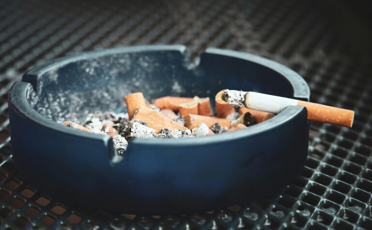 Popelník plný nedopalků s hořící cigaretou položenou na okraji stolku.