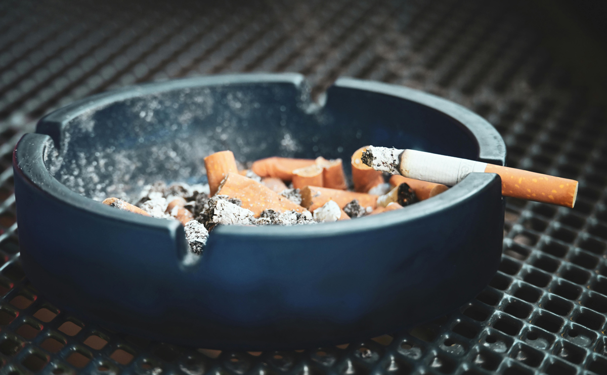 Popelník plný nedopalků s hořící cigaretou položenou na okraji stolku.