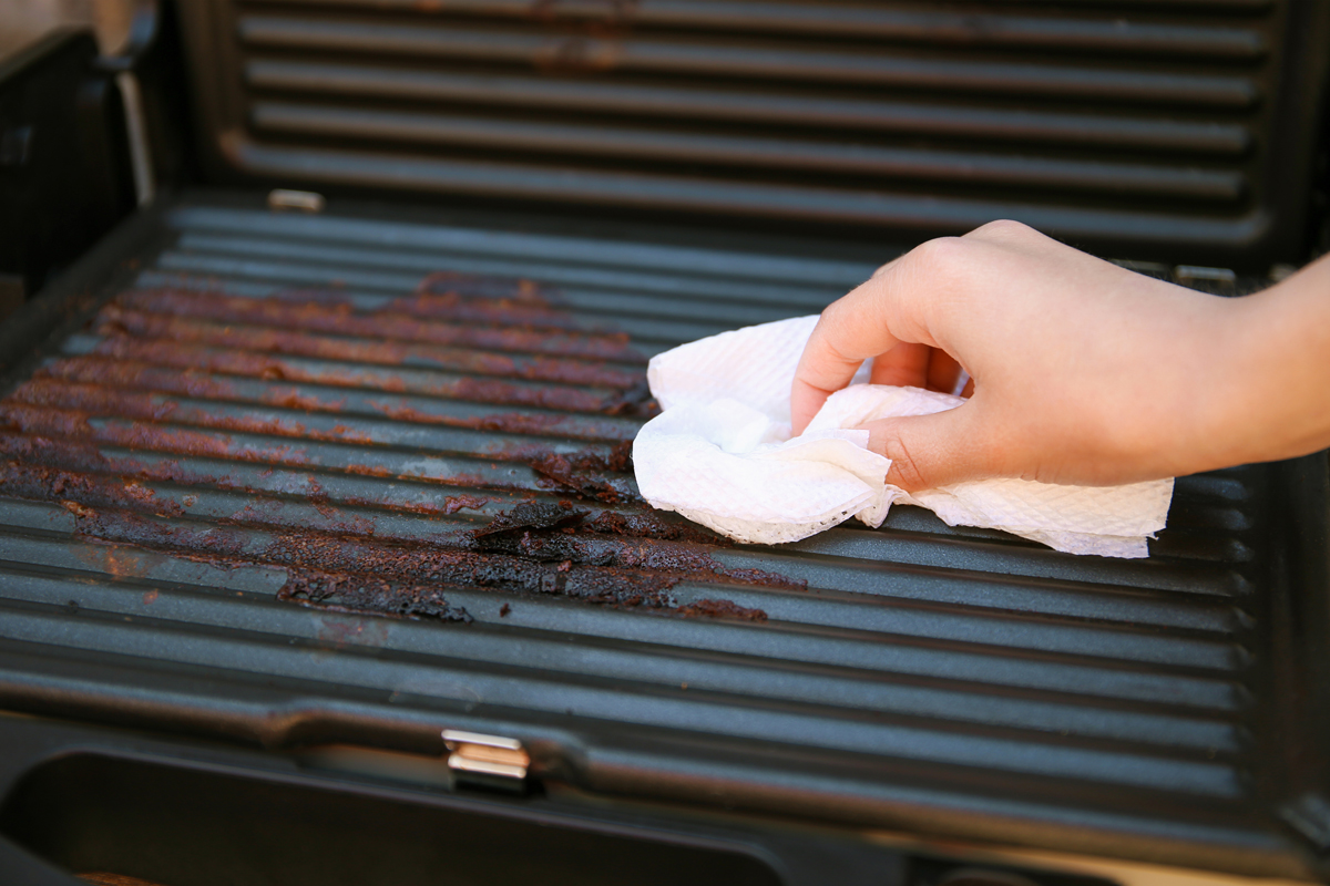 Ruka čistí grilovací rošt elektrického grilu papírovým ubrouskem.