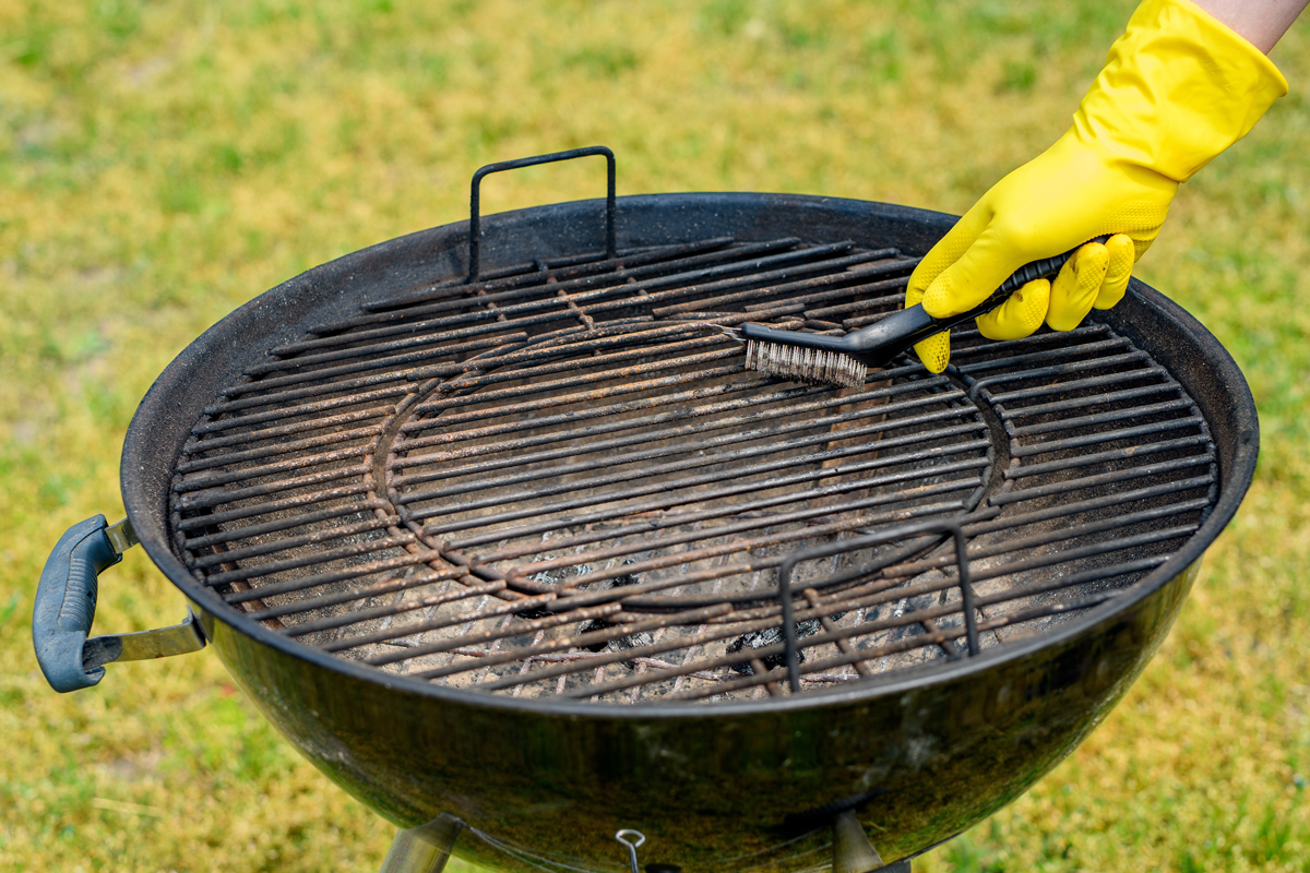 Ruka v žluté rukavici čistí kulatý rošt kotlového grilu drátěným kartáčem; po dopečení zavřete víko, nechte zbytky 10–15 min vyhořet, ještě teplý rošt vykartáčujte po směru prutů, vysypte popel a rošt lehce naolejujte (pozor na uvolněné štětiny).