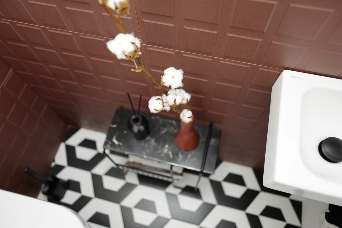 Detail toalety s 3D obkladem v hnědém odstínu kakaa, černobílou grafickou dlažbou a mramorovou konzolí s vázami a větvičkou bavlníku; atmosféra elegantní, kontrastní a čistá.