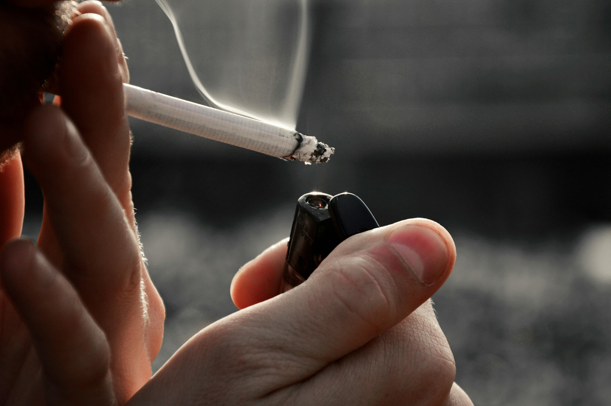 Detail rukou zapalujících cigaretu zapalovačem, z cigarety se zvedá kouř na tmavém pozadí.