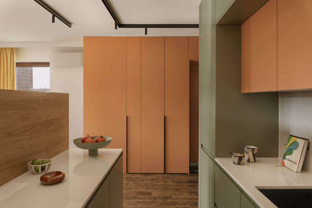 Moderní kuchyň s matnými skříněmi v šalvějově zelené a terakotově oranžové, bílými deskami, dřevěným paravánem a kolejnicovým osvětlením.