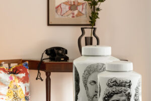 Lesklá deska stolu se dvěma porcelánovými dózami s antickými hlavami; v pozadí konzolový stolek s černým rotačním telefonem, váza s větvičkou a zarámovaný obraz klauna.