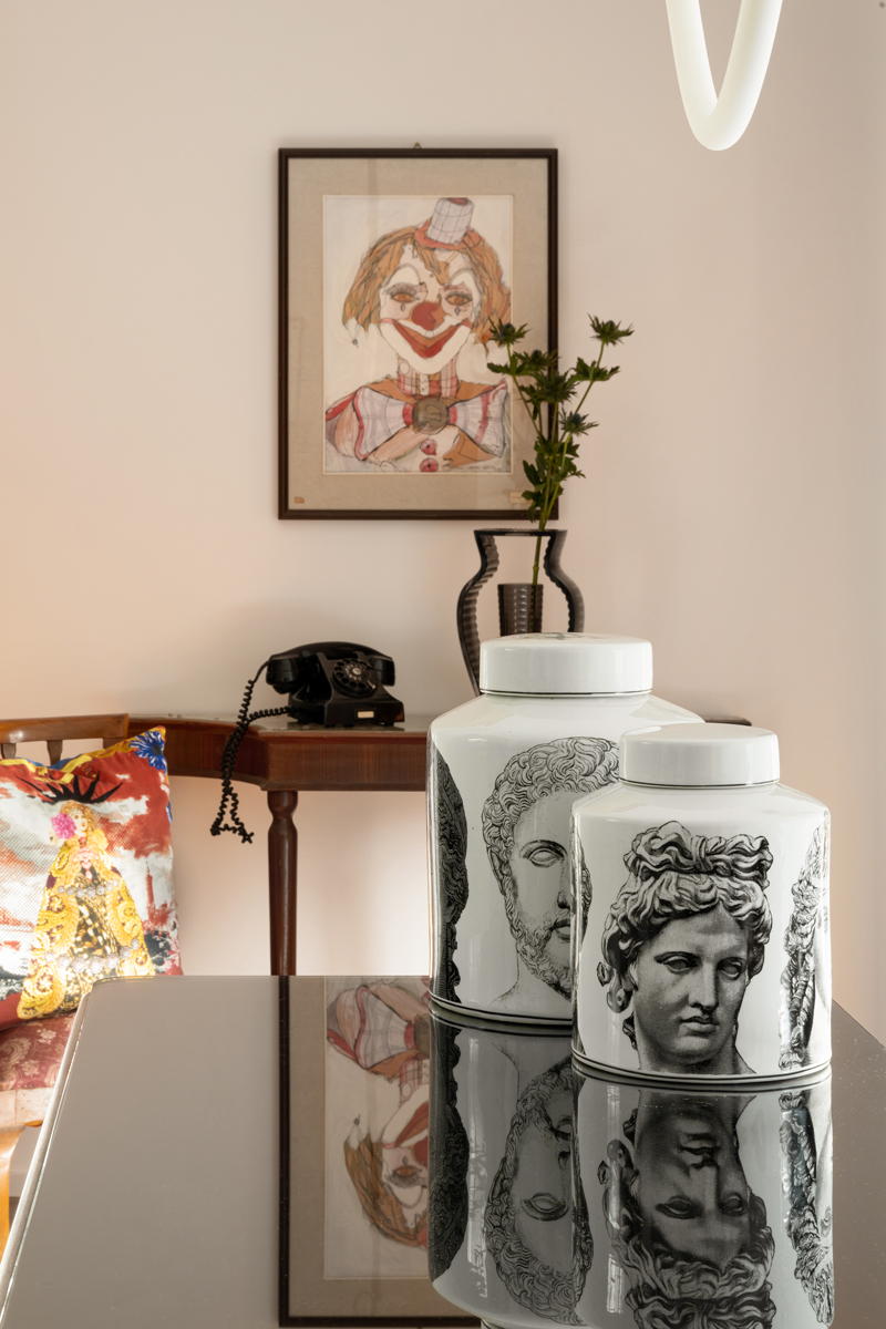 Lesklá deska stolu se dvěma porcelánovými dózami s antickými hlavami; v pozadí konzolový stolek s černým rotačním telefonem, váza s větvičkou a zarámovaný obraz klauna.