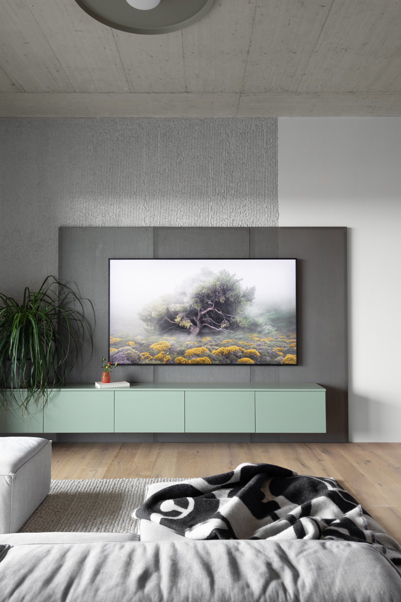 Obývací stěna s velkou TV na šedém panelu, pod ní závěsná skříňka v mátové barvě; před ní šedá sedačka a tkaný koberec.