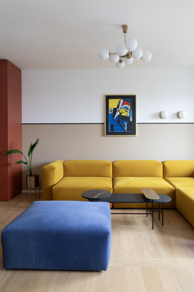 Moderní obývák s hořčicovou modulární sedačkou, kobaltovým pufem a grafickým obrazem; hravá, galerijní atmosféra s teplými neutrály.