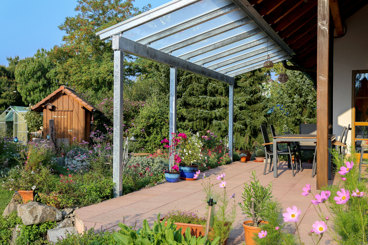 Zahradní pergola s pozinkovanou konstrukcí a průhlednou střechou na dlažbě, kolem kvetoucí záhony, zahradní domek a skleník.