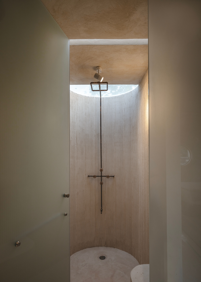 Minimalistická sprcha s kovovou hlavicí a kruhovým stropním oknem, které přivádí denní světlo do prostoru s jemně rýhovaným obkladem.