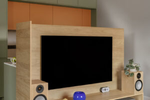 Samostatná TV stěna z dubu s velkou obrazovkou, bočními reprobednami, červenými zásuvkami a policí s elektronikou a knihami; dekor tvoří modrá váza a rostlina; za ní je vidět kuchyň v olivově zelené a terakotě, nad stropem černé lištové světlo, na zemi tmavá dřevěná podlaha.