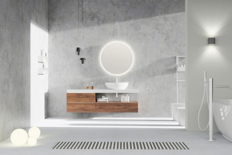 Elegantní koupelna s betonovými stěnami, moderním dřevěným umyvadlovým pultem a kruhovým podsvíceným zrcadlem.