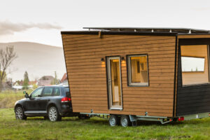 Dům na kolečkách tažený autem po louce, dřevěný obklad, velká okna a střešní solární panely; večerní světlo.