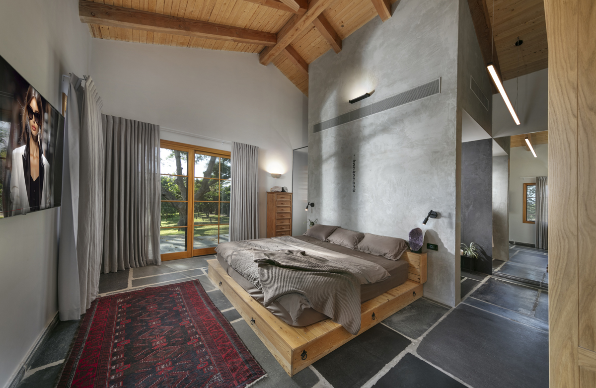 Designová ložnice s postelí z masivního dřeva, šedými betonovými stěnami, orientálním kobercem a výhledem do zahrady přes francouzská okna.