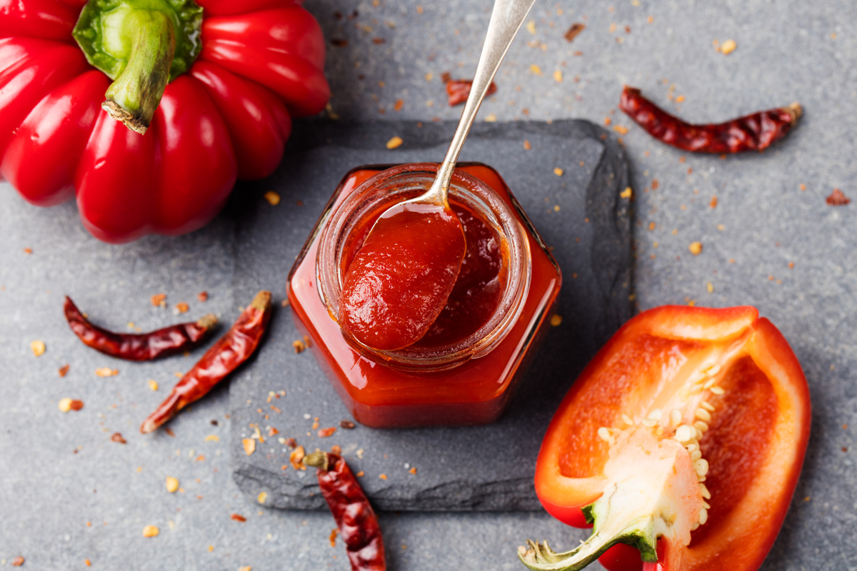 Domácí paprikový a chilli džem v malé sklenici s čerstvou i sušenou paprikou kolem.